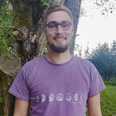 Auf dem Bild ist das Vorstandsmitglied Lukas Ermann. Er steht vor einem Baum, trägt eine Brille und lacht in die Kamera.
