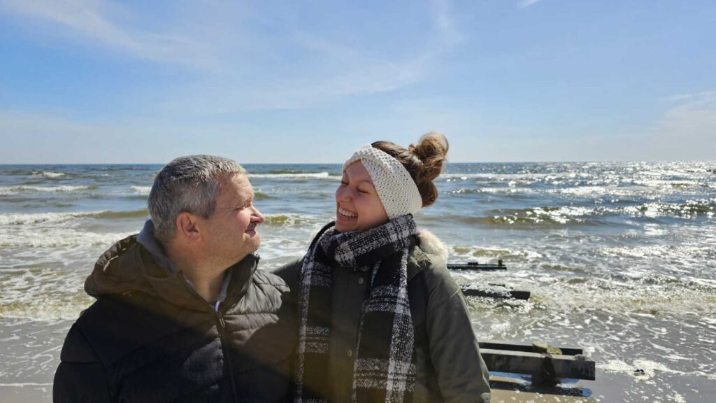 Eine Reisebegleiterin mit Strickstirnband und kariertem Schal lacht herzlich und schaut einen Mann an, der zurücklächelt. Beide stehen in Winterjacken am sonnigen Strand, dahinter Wellen und Holzbuhnen im glitzernden Meer.