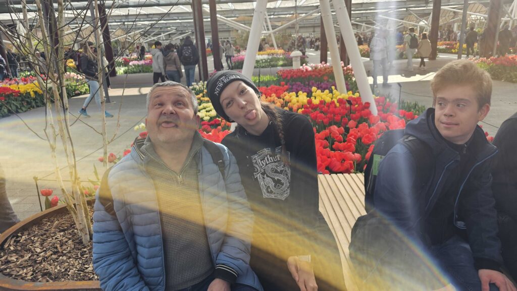 Eine Reisebegleiterin sitzt neben einem Reisenden auf einer Holzbank. SIe strecken beide die Zunge heraus. Hinter ihnen blühen rote, gelbe und lila Tulpen.