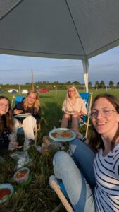 4 Personen machen auf dem Hurricane Camping platz ein Selfie. Alle 4 sitzen auf Strandstühlen auf einer Wiese und haben Teller mit Essen in der Hand. Alle schauen fröhlich in die Kamera und die Sonne scheint.