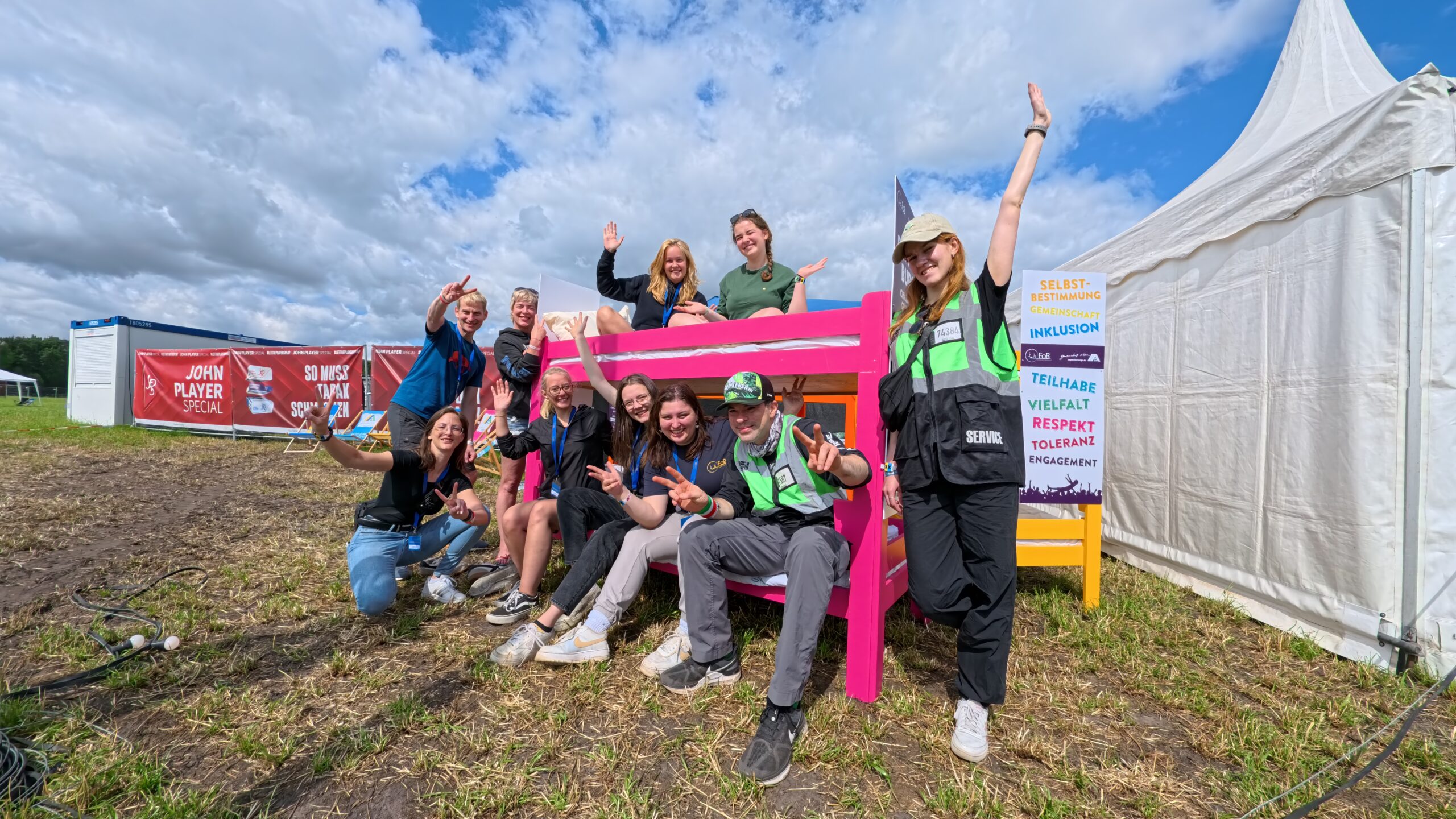 Gruppenfoto des FoB-Teams vor dem Special Needs Camp auf dem Hurricane Festival 2025. Die Gruppe sitzt und steht lachend um ein pinkes Etagenbett. Im Hintergrund ein weißes Zelt und ein bunter Aufsteller mit Begriffen wie Inklusion, Vielfalt und Respekt. Alle wirken fröhlich und motiviert.