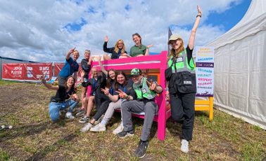 Gruppenfoto des FoB-Teams vor dem Special Needs Camp auf dem Hurricane Festival 2025. Die Gruppe sitzt und steht lachend um ein pinkes Etagenbett. Im Hintergrund ein weißes Zelt und ein bunter Aufsteller mit Begriffen wie Inklusion, Vielfalt und Respekt. Alle wirken fröhlich und motiviert.