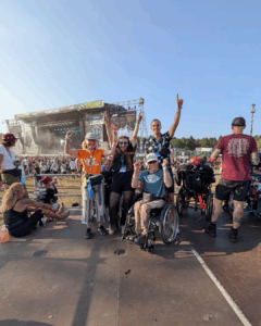 Es sind einige Mneschen auf dem Festivalgelände zu sehen. Alle sind sehr föhlich. Ein paar Menschen sitzen im Rollstuhl. Alle jubeln föhlich in die Kamera. Im Hintergrund ist eine Bühne zu sehen.