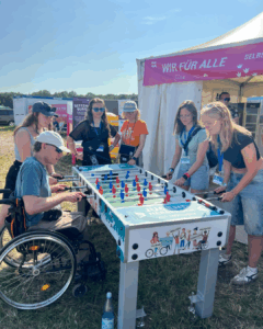 6 Leute stehen um einen Kicker rum. 4 spielen am Kicker. Eine Person sizt im Rollstuhl. Die Sonne scheint.