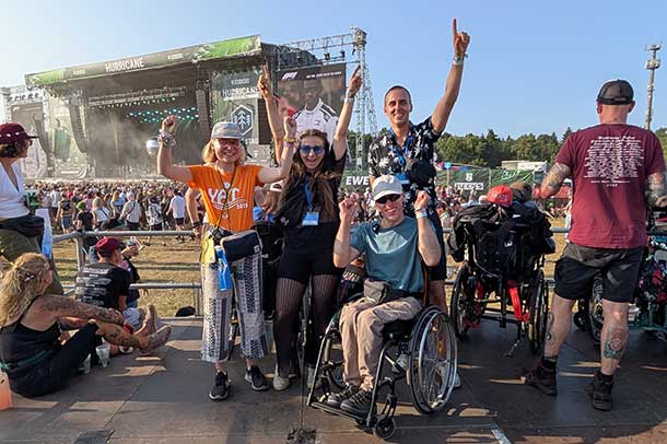 Eine Gruppe von vier Personen blickt in die Kamera. Sie freuen sich und haben jubelnd die Hände in die Luft gehoben. Eine Person sitzt im Rollstuhl. Im Hintergrund sind ein Festivalgelände und eine Bühne zu sehen.