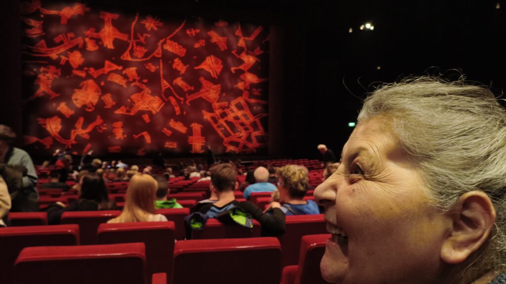 Rechts im Bild sieht man das Gesicht einer Frau von der Seite. SIe sitzt in einem Musical Saal mit roten Stühlen und lacht.