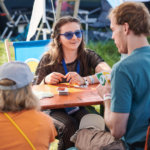Junge Frau sitzt an einem Tisch und blickt zwei Menschen an, die mit dem Rücken zur Kamera sitzen.