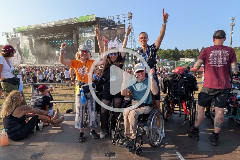 Eine Gruppe von vier Personen blickt in die Kamera. Sie freuen sich und haben jubelnd die Hände in die Luft gehoben. Eine Person sitzt im Rollstuhl. Im Hintergrund sind ein Festivalgelände und eine Bühne zu sehen. Über den Personen ist eine Zusatzgrafik eingeblendet, die ein Video markiert.