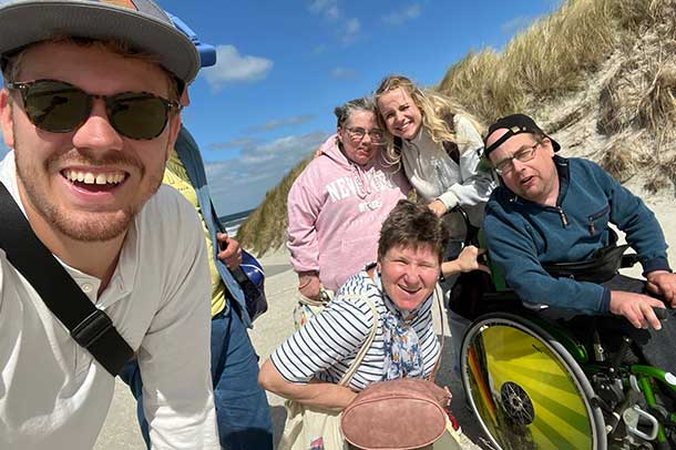 Peers Eine Gruppe von 5 Personen macht ein Selfie am Strand. Alle lachen in die Kamera. Eine der Personen sitzt im Rollstuhl. Im Hintergrund sind der blaue Himmel und Dünen zu sehen.