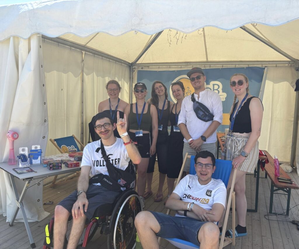 Das FoB-Team posiert gemeinsam am Infostand unter einem weißen Zelt auf einem Festival. Sieben Personen lächeln in die Kamera, darunter eine Person im Rollstuhl im Vordergrund. Im Hintergrund ist das FoB-Banner zu sehen, auf dem Tisch liegen Infomaterialien. Die Stimmung ist locker und herzlich.