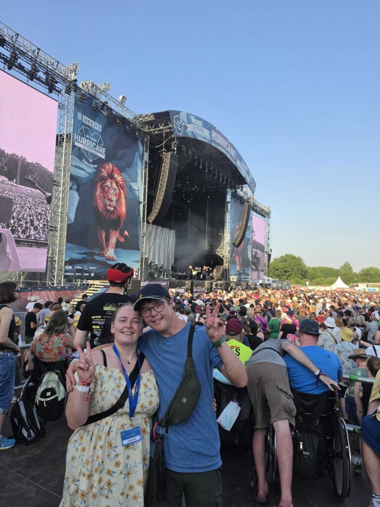 Zwei junge Menschen lächeln und zeigen das Peace-Zeichen vor der Hauptbühne des Hurricane Festivals. Rechts daneben ist eine Person im Rollstuhl zu sehen. Im Hintergrund eine große Menschenmenge und die Festivalbühne bei strahlendem Sonnenschein