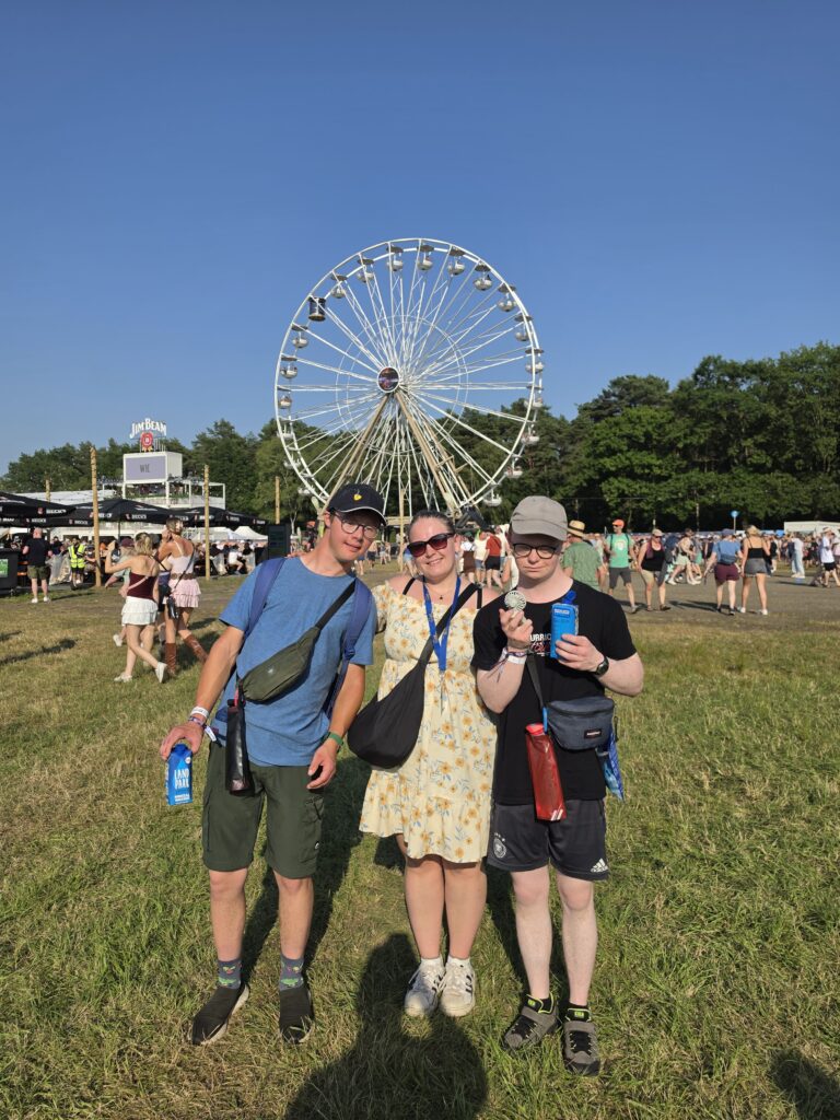 Drei Personen posieren auf einem Festivalgelände vor einem Riesenrad.