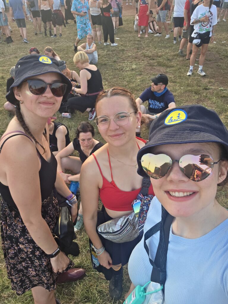 Drei junge Frauen lächeln in die Kamera bei einem Outdoor-Festival. Zwei von ihnen tragen Bucket Hats mit dem FoB-Logo. Im Hintergrund sitzen und stehen viele Festivalbesucher auf einer Wiese bei warmem Abendlicht.