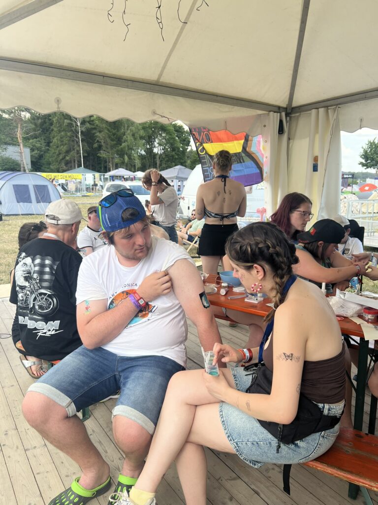 Eine junge Frau mit FoB-Lanyard klebt einem Mann am Tisch ein Tattoo auf den Arm. Um sie herum sitzen weitere Festivalbesucher unter einem weißen Zelt. Im Hintergrund ist eine Progress-Pride-Flagge zu sehen sowie das Festivalgelände mit Zelten und Bäumen.