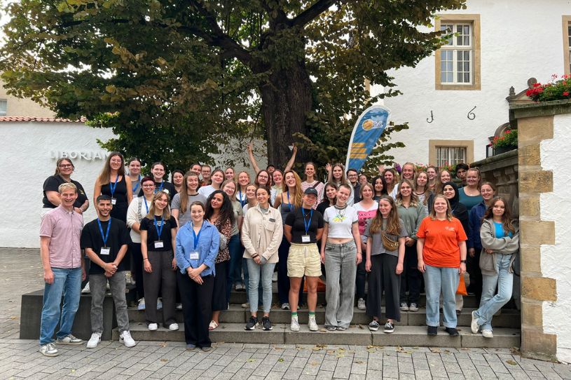 Das Foto zeigt eine große Gruppe an Personen, die gemeinsam für die Kamera posieren. Im Hintergrund sind ein Haus und ein Baum zu sehen.