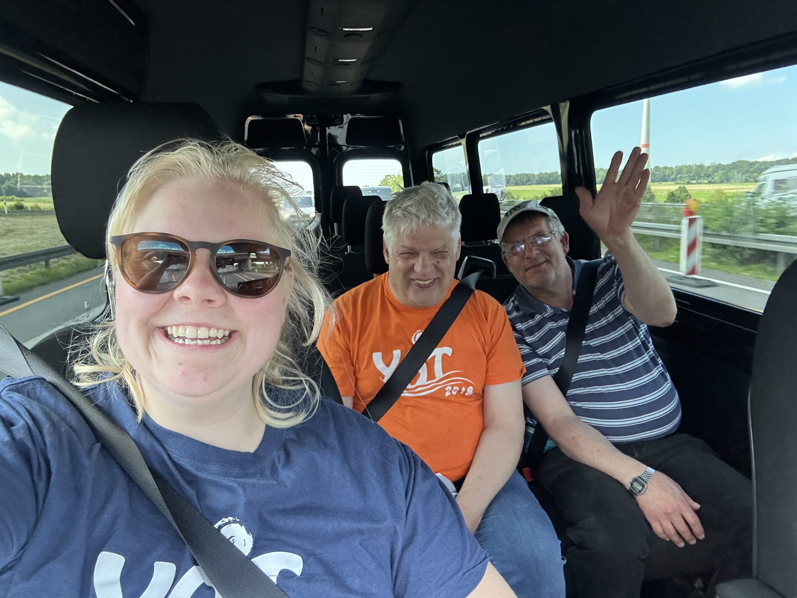 Selfie einer lachenden Reisebegleiterin mit zwei männlichen Teilnehmern im YAT-Bus während der Fahrt, einer trägt ein oranges YAT-2018-Shirt, der andere winkt fröhlich in die Kamera.