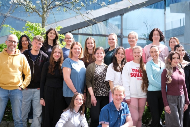 Gruppenfoto eines Teams von etwa 18 Personen vor einer modernen Glasfassade, aufgestellt in mehreren Reihen im Freien bei Sonnenschein, im Hintergrund ein blühender Baum.