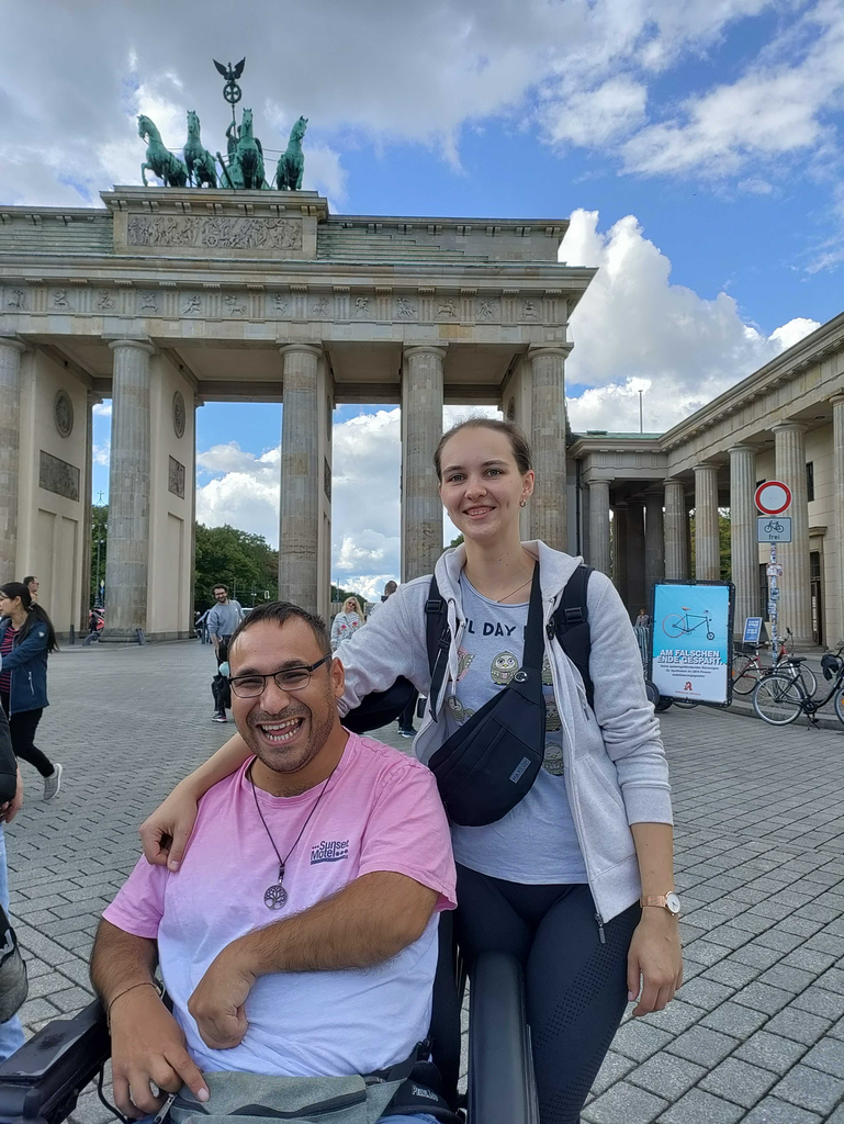 Reisebegleiterin legt den Arm um einen strahlenden Reisegast im Rollstuhl. Hinter ihnen sieht man das Brandenburger Tor in Berlin. Der Himmel ist blau mit Wolken.